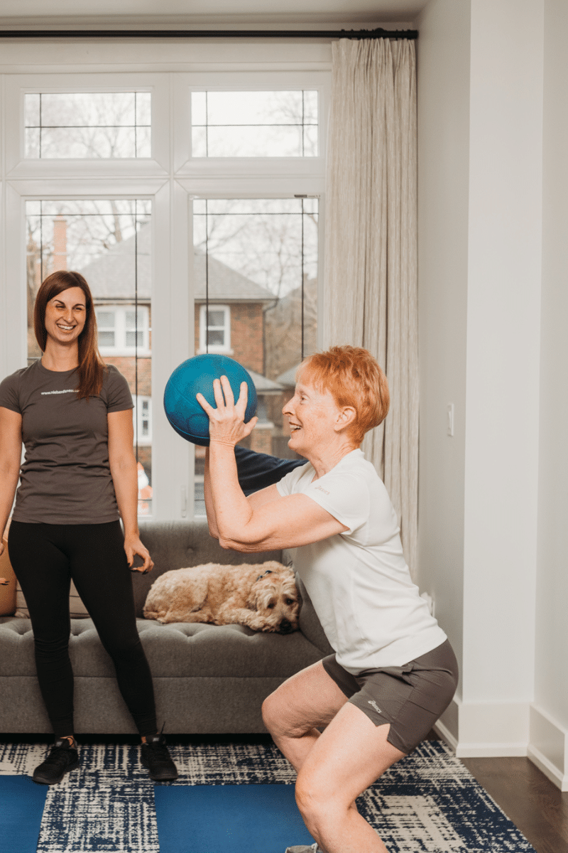 woman holds medicine ball and squats while a trainer observes